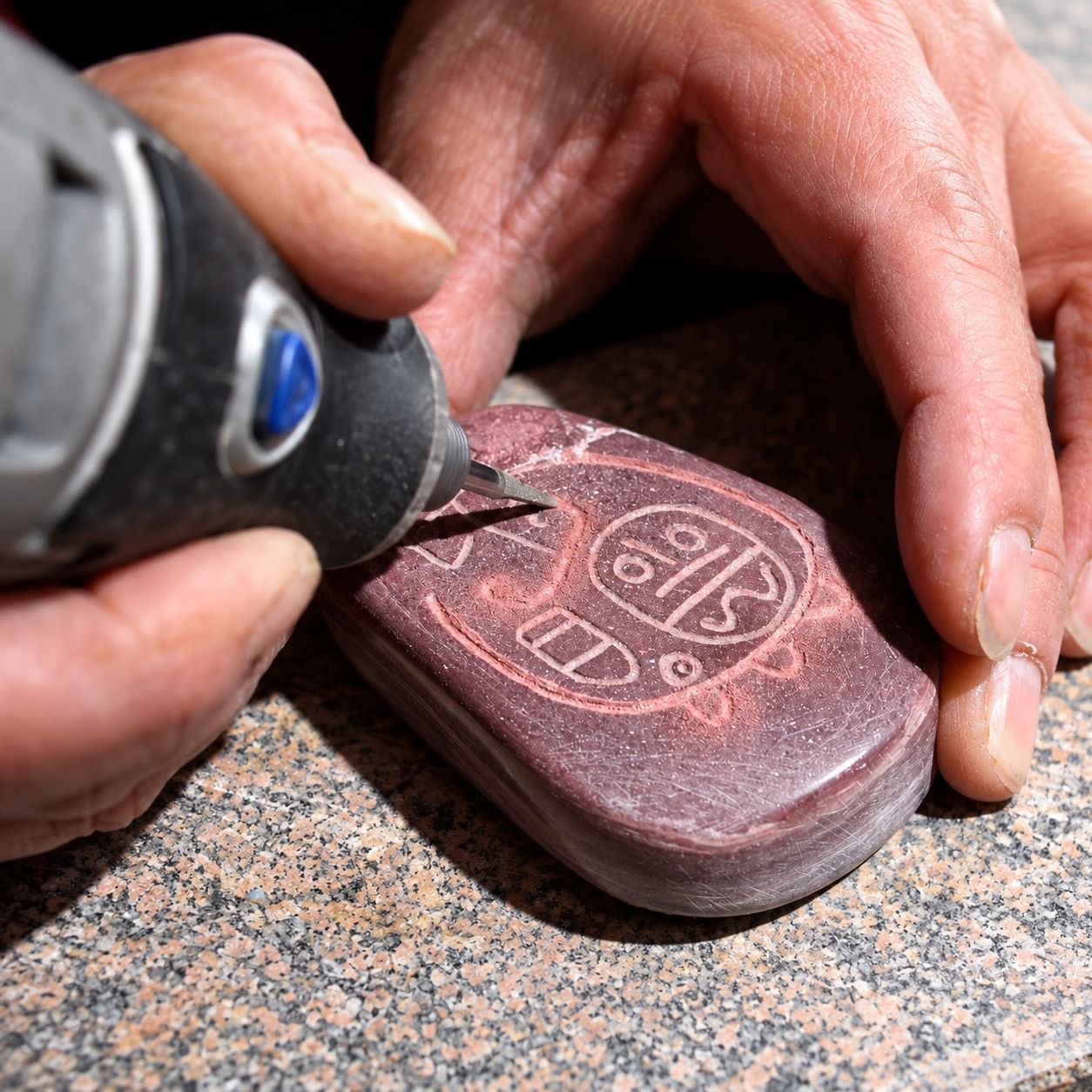 Manos grabando con una herramienta rotativa un diseño de líneas sobre una piedra rojiza pulida.